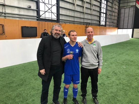 Standing is south Melbourne FC player Brendan Spencer holding his award. To his right standing is Dave Connolly, National manager of Australian Blind Football and to his left Kimon Taliadoros, CEO of Football Victoria